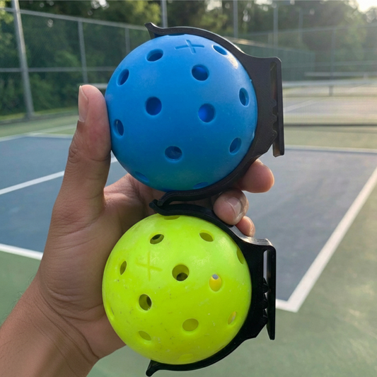 Hand holding two pickleballs in a holder on a tennis court